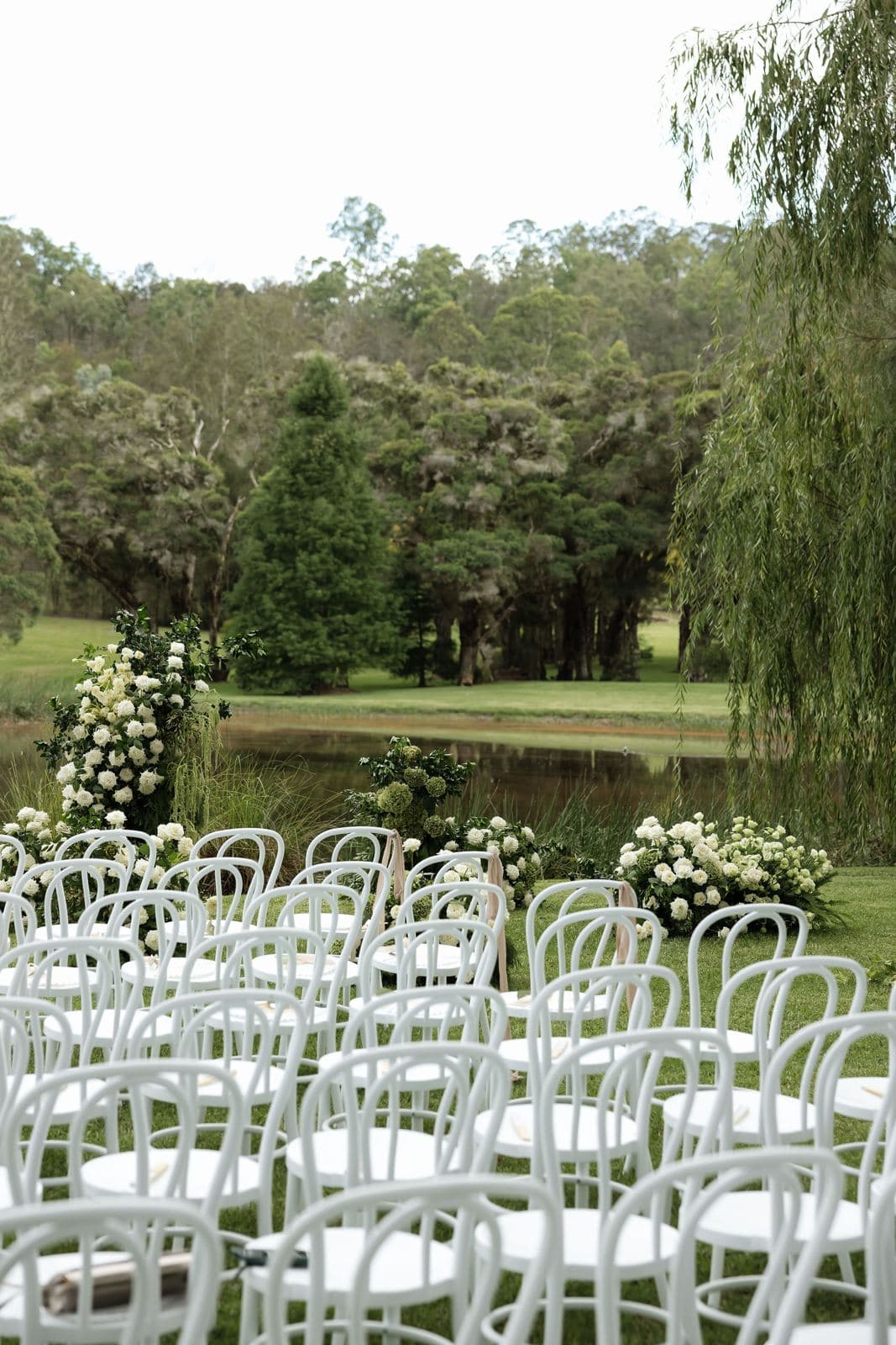 White bentwood ceremony chairs beside a willow-lined lake
